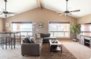 Amplia sala de estar con un sofá seccional grande, una mesa de centro y un comedor con mesa y sillas. Dos ventanas ofrecen luz natural y vistas al exterior.