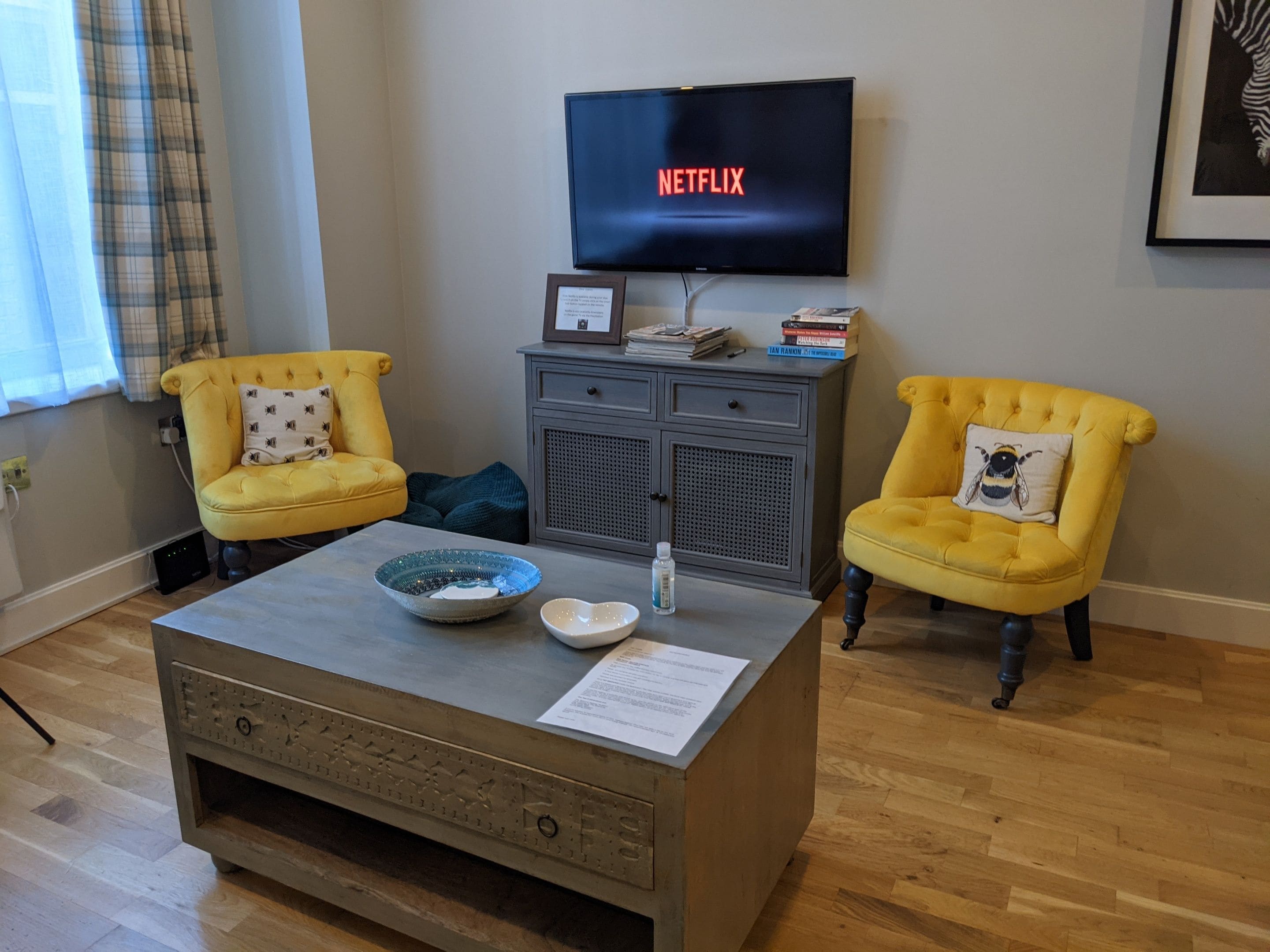 A living room with two yellow armchairs, a coffee table with a bowl and a bottle, a grey cabinet, a television displaying 'NETFLIX', and a framed picture on the cabinet.