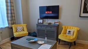 Ett vardagsrum med en tv-skärm som visar "NETFLIX", två gula fåtöljer med kuddar, ett soffbord med en skål och en flaska, och ett grått skåp med böcker.