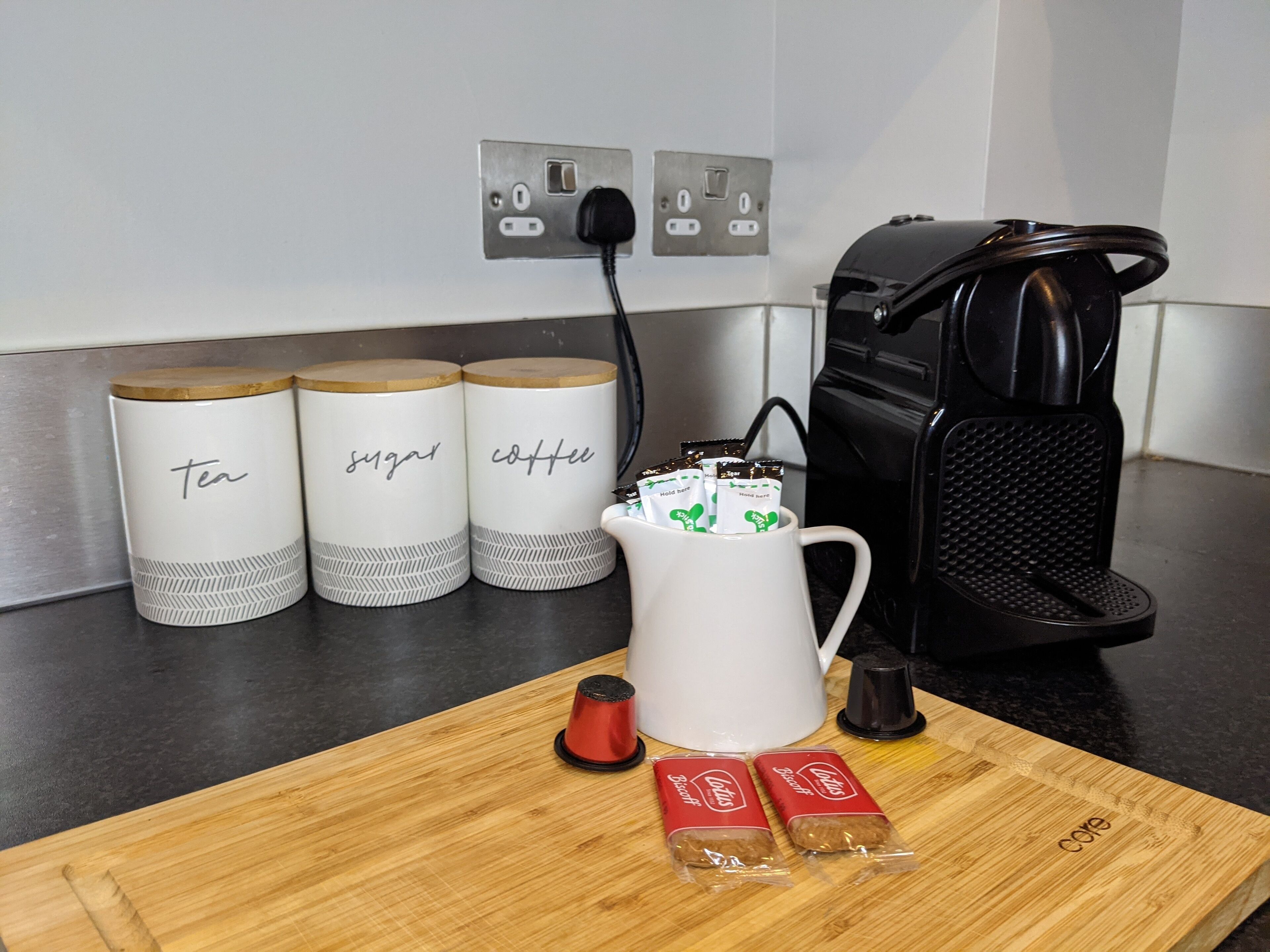 Una encimera de cocina con una cafetera, una taza con cápsulas de café y dos recipientes etiquetados como 'té' y 'café'.