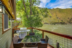 A balcony with a grill, wicker furniture, and a view of a river and hills. - Cabin on the Salmon River (Riggins)