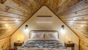 A cozy attic bedroom with a sloped ceiling, a bed with a patterned duvet, two bedside tables with lamps, and a wall-mounted air conditioning unit.