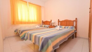 A room with two beds, each with a striped bedspread, a nightstand with a lamp, and a window with yellow curtains. - Apartamentos Marblau Las Alondras (Gandia)