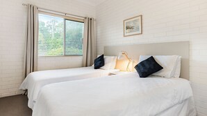 Een hotelkamer met twee bedden, een raam met luxaflex, een bedlampje en een ingelijst schilderij aan de muur.