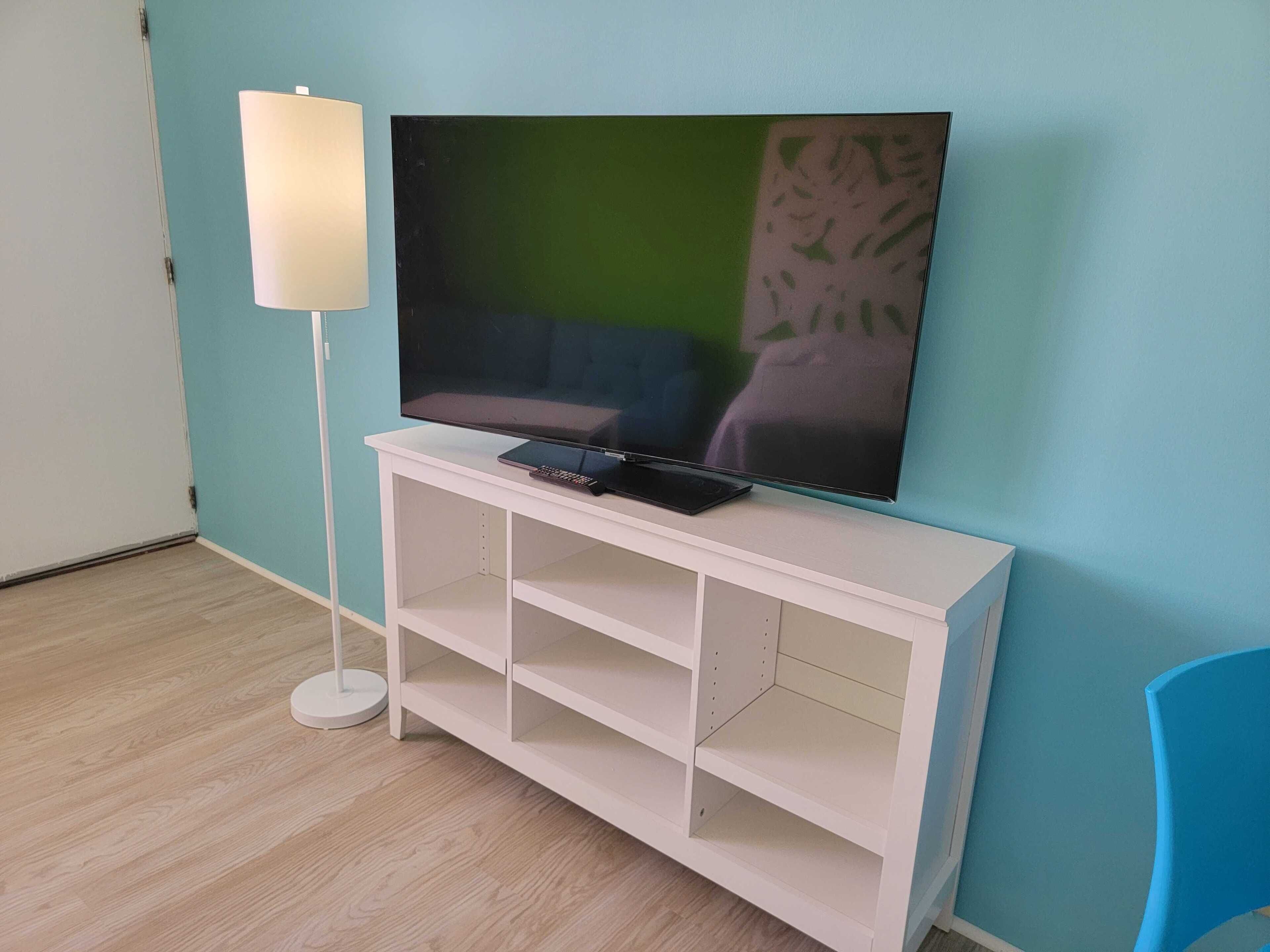A room with a flat-screen TV on a white stand, a floor lamp, and a blue chair.