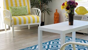 Um quarto com uma cadeira de cor branca e listras, uma mesa pequena, um tapete azul estampado, uma planta em vaso e uma janela com vista.