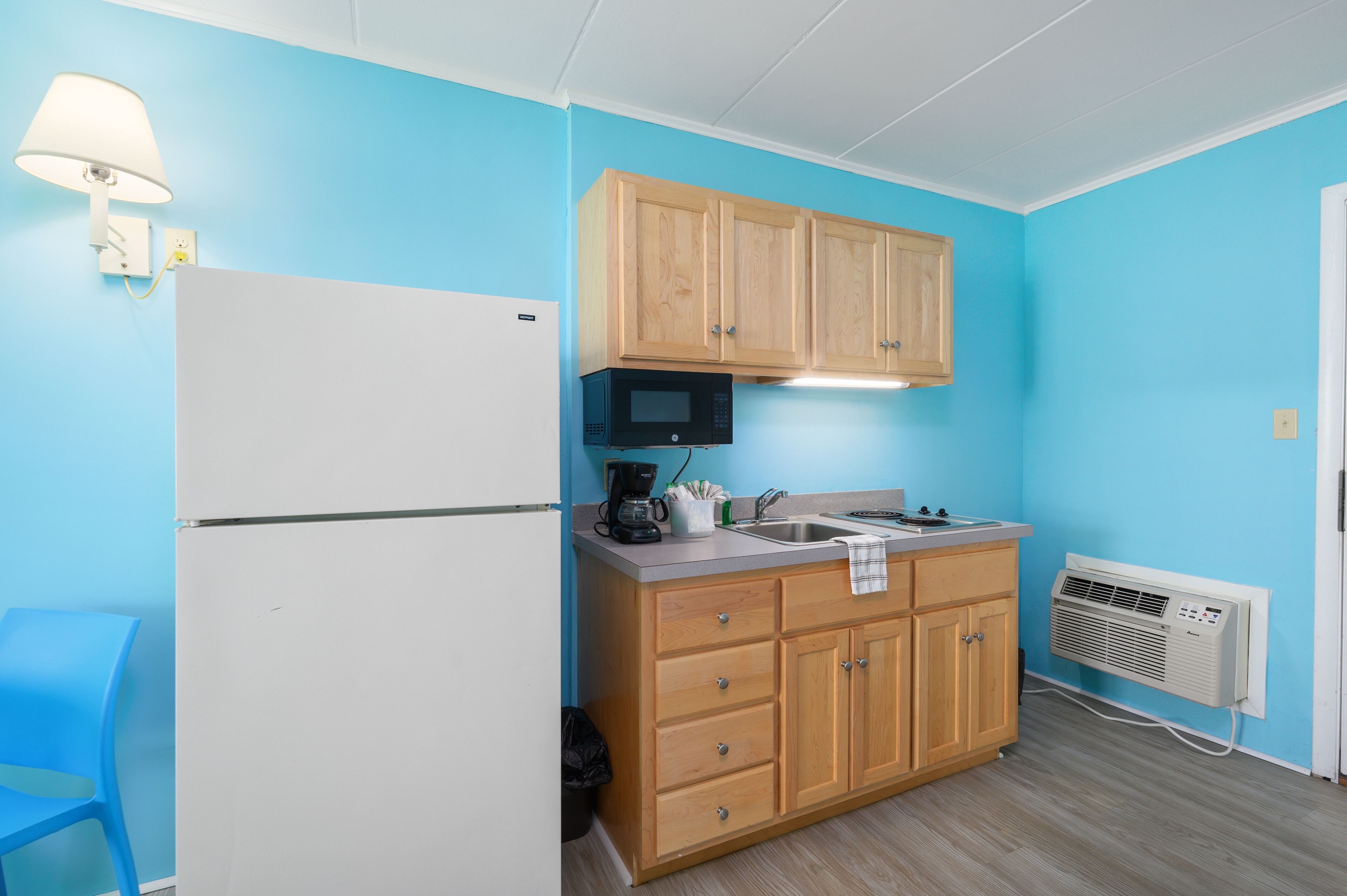 A compact kitchen with a white refrigerator, wooden cabinets, a microwave, and a built-in air conditioner.