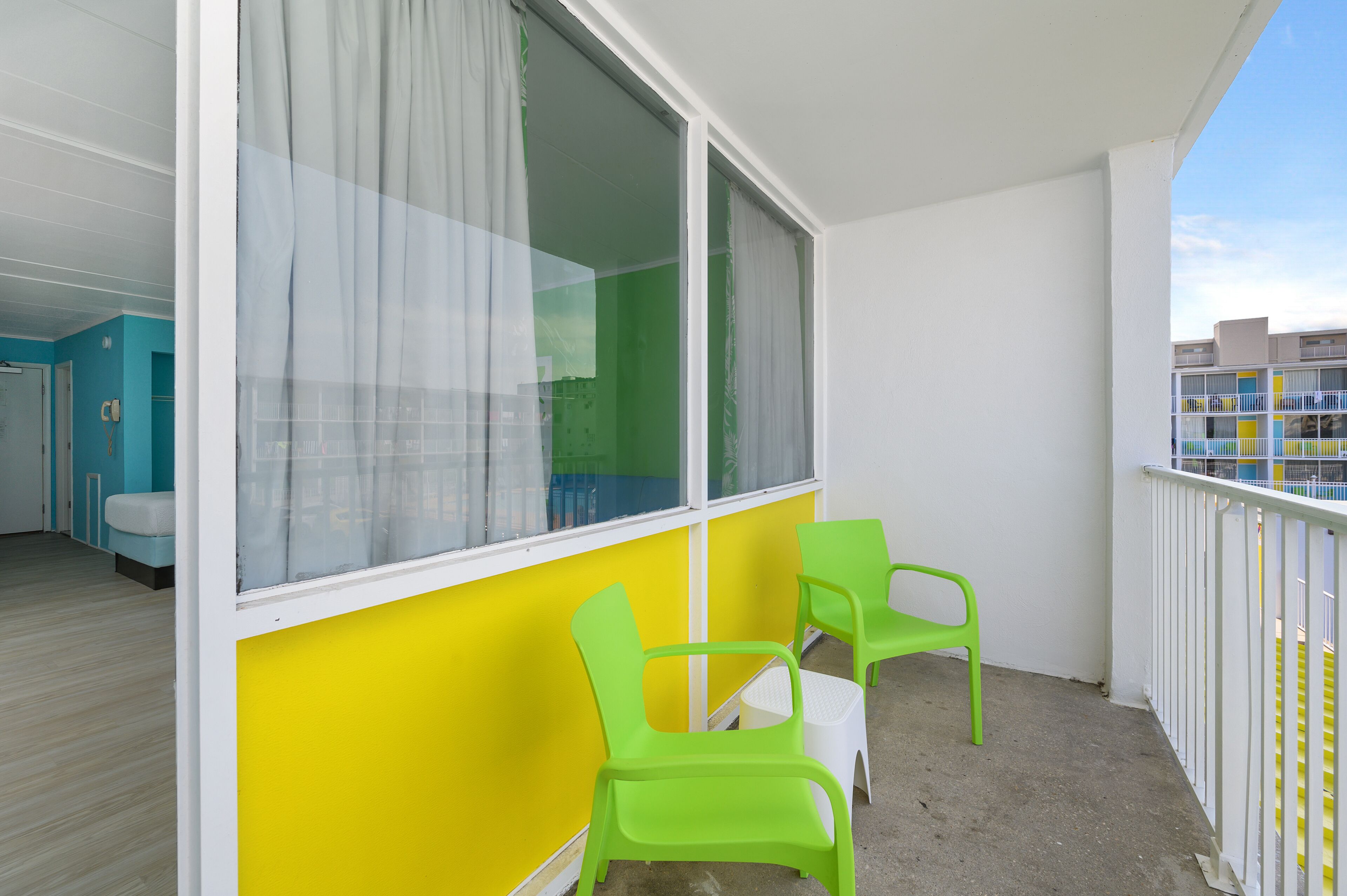 Un balcón con dos sillas verdes y una mesita, cortinas blancas en las ventanas, y vistas a un edificio con balcones.
