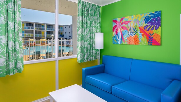 Quarto com sofá azul, mesa branca, pintura abstrata colorida, e vista para uma piscina e um edifício através de cortinas verdes.