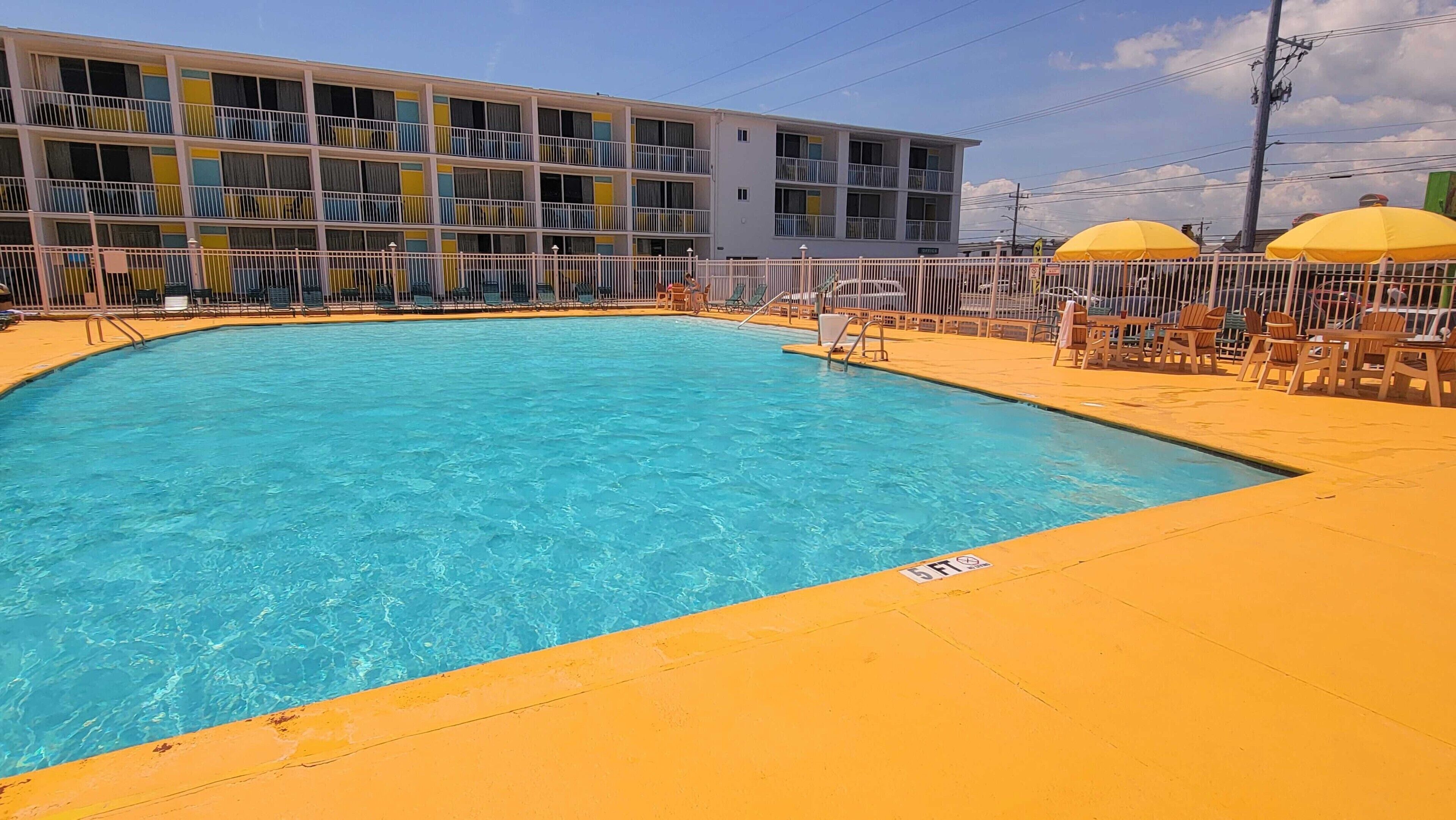 Una piscina con un borde amarillo, rodeada por una valla blanca y un edificio con balcones.
