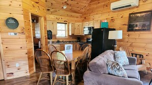 Interior de una cabaña con paredes de madera, un comedor con sillas de madera, un sofá púrpura y una cocina con refrigerador negro.