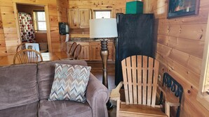 Un interior de cabaña acogedor, con una mesa de comedor de madera, un sofá con cojines estampados, una silla de madera y una zona de cocina con armarios y un refrigerador.