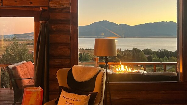A cozy cabin interior with a large window overlooking a lake and mountains. A chair with a fur throw and a pillow labeled "Hennys Lake Idaho" is positioned near the window. A table lamp and a fire pit are also visible. - NO BETTER VIEW THAN THIS (Island Park)