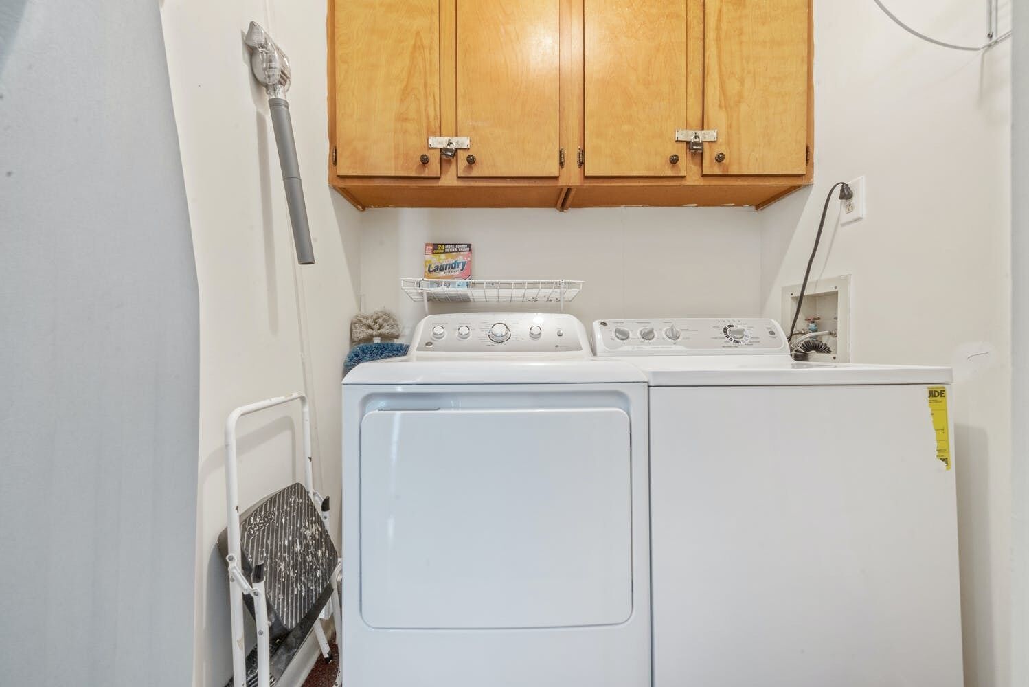 A laundry area with a washer and dryer, wooden cabinets above, and a metal rack with a laundry detergent box.