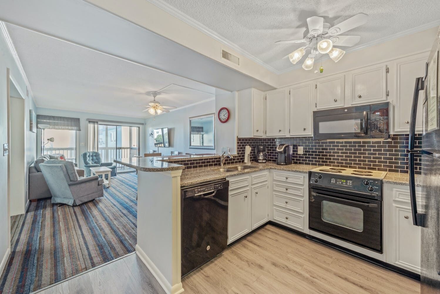 A modern kitchen with white cabinets, a black refrigerator, and a microwave. There is a dining area with a gray sofa and a striped rug.