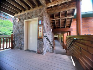 A wooden staircase leading to a door with a stone wall, a wooden railing, and a view of a building in the background.
