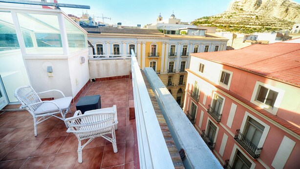 Un balcon agrémenté de fauteuils et d’une table en osier, offrant une vue sur le paysage urbain, avec ses bâtiments historiques et une colline.
