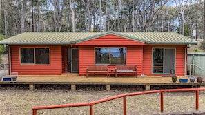 A red wooden house with a green roof, a porch with a bench, and a fenced yard.