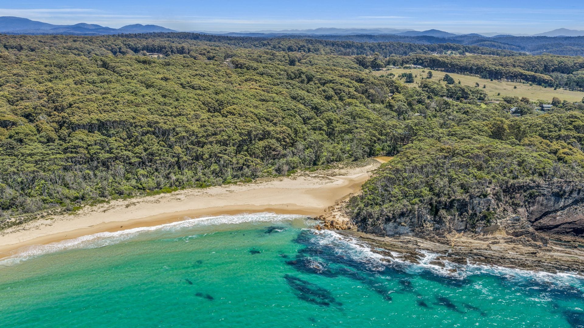 An aerial view of a coastal area with a beach, clear turquoise waters, and dense forested hills.