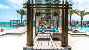 Zona de piscina con un pérgola de madera, sillas de descanso y vistas al océano y edificios de gran altura.
