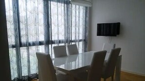 Un comedor con una mesa rectangular y seis sillas, un televisor de pantalla plana montado en la pared, y cortinas con un estampado.