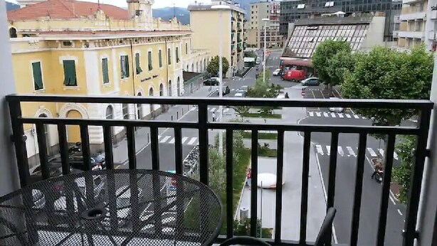 Un balcón con una mesa y sillas de metal, con vistas a una calle con edificios y una panorámica despejada del entorno.