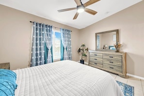 Une chambre à coucher avec un lit, un ventilateur de plafond, une commode et une fenêtre avec des rideaux.