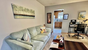 Un salón con un sofá gris, una mesa de centro con libros y un control remoto, un cuadro en la pared, una cocina al fondo y un escritorio con una silla.