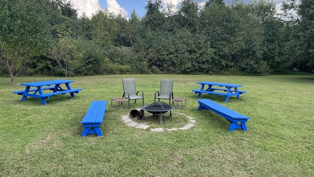 Un espacio al aire libre con césped, mesas y sillas de picnic, una fogata y un bosque denso al fondo.