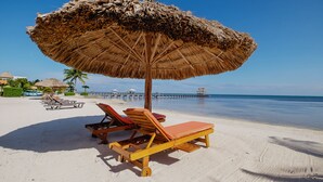 Une plage avec des chaises longues en bois et un parasol en chaume, un ciel bleu dégagé et une jetée en arrière-plan.