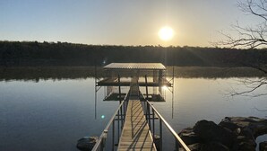 A wooden pier leading to a metal-roofed dock on calm water, surrounded by rocks and trees under a clear sky. - Waterfront house with private dock and boat slip (Stigler)