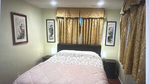 A bedroom with a bed, two bedside tables, a nightstand, a window with curtains, and framed artwork on the walls.