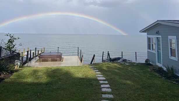 Une zone herbeuse avec un sentier de pierres menant à une jetée surplombant l’eau, une petite maison et un arc-en-ciel dans le ciel.