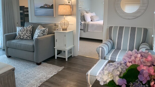 Una sala de estar moderna con un sofá gris, un sillón a rayas y una mesita de noche blanca con una lámpara. Se ve una cama con cabecera estampada al fondo.