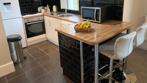 A modern kitchen with white cabinets, a black backsplash, a microwave, and a fruit basket on the counter. - Elegant 2 Bed Georgian Apartment in Herne Bay (Herne Bay)