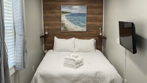 A neatly made bed with a white comforter and pillows, a painting of a beach scene on the wall, and a television mounted on the wall. - Water Front Home: Fisherman's' dream with private dock and beautiful sunsets (Brownstown Charter Township)