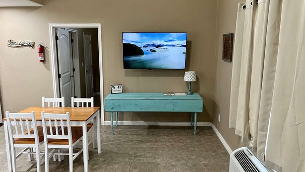 A room with a wooden table and white chairs, a teal console table with a lamp, a TV on the wall, and a fire extinguisher. - Hunter's Ridge Cabins - 1 (Concan)
