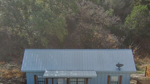 A small, single-story building with a metal roof, a porch with two chairs, and a satellite dish. - Hunter's Ridge Cabins - 1 (Concan)