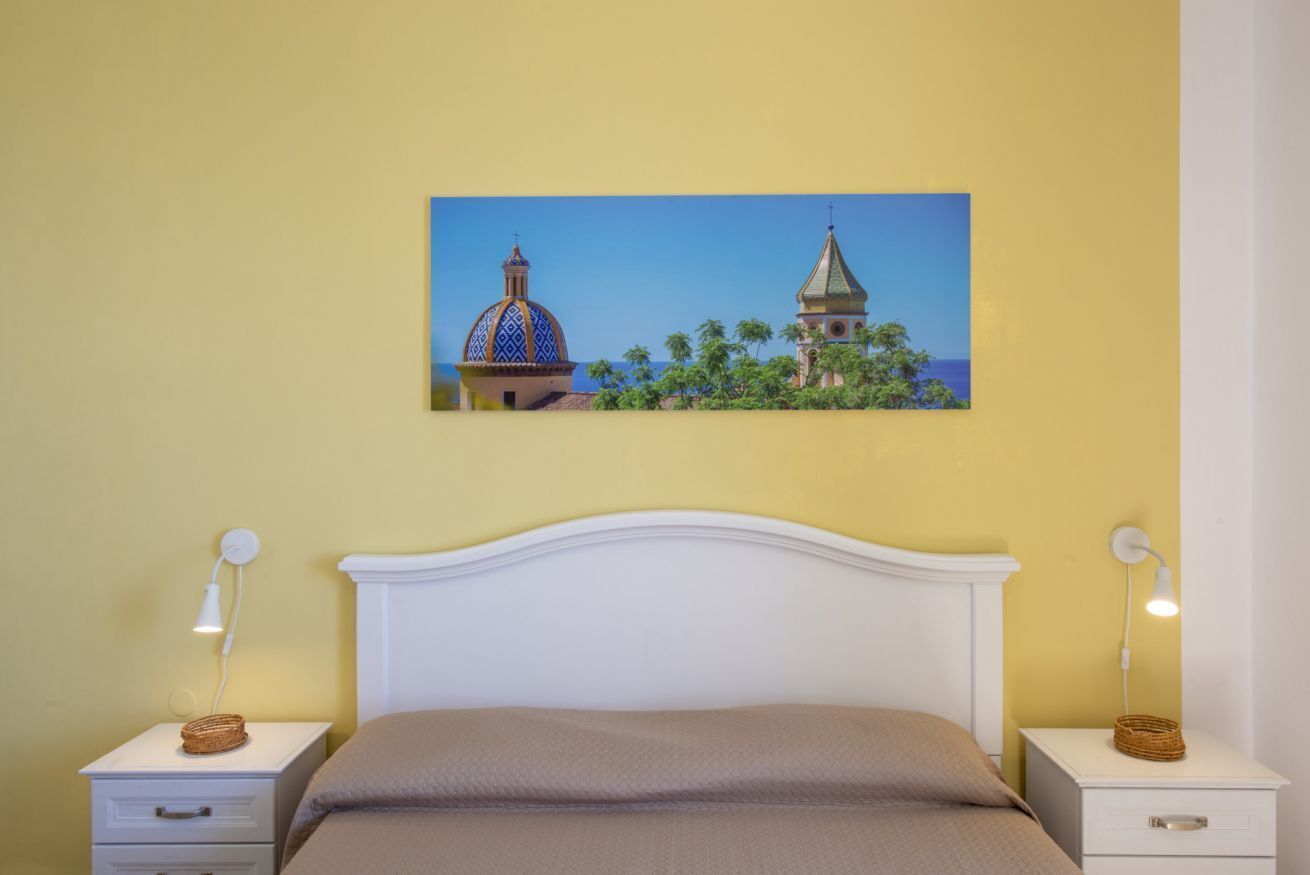 A bedroom with a white bed, two bedside tables with lamps, a painting of a building and trees on the wall, and a yellow wall.