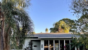 A modern two-story house with a covered patio, surrounded by lush greenery and a clear blue sky.