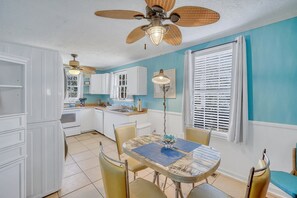 A kitchen with white cabinets, a dining table with chairs, a ceiling fan, and a window with blinds. - Tybee Lighthouse Cottage (B): Walk to the Beach! (Tybee Island)