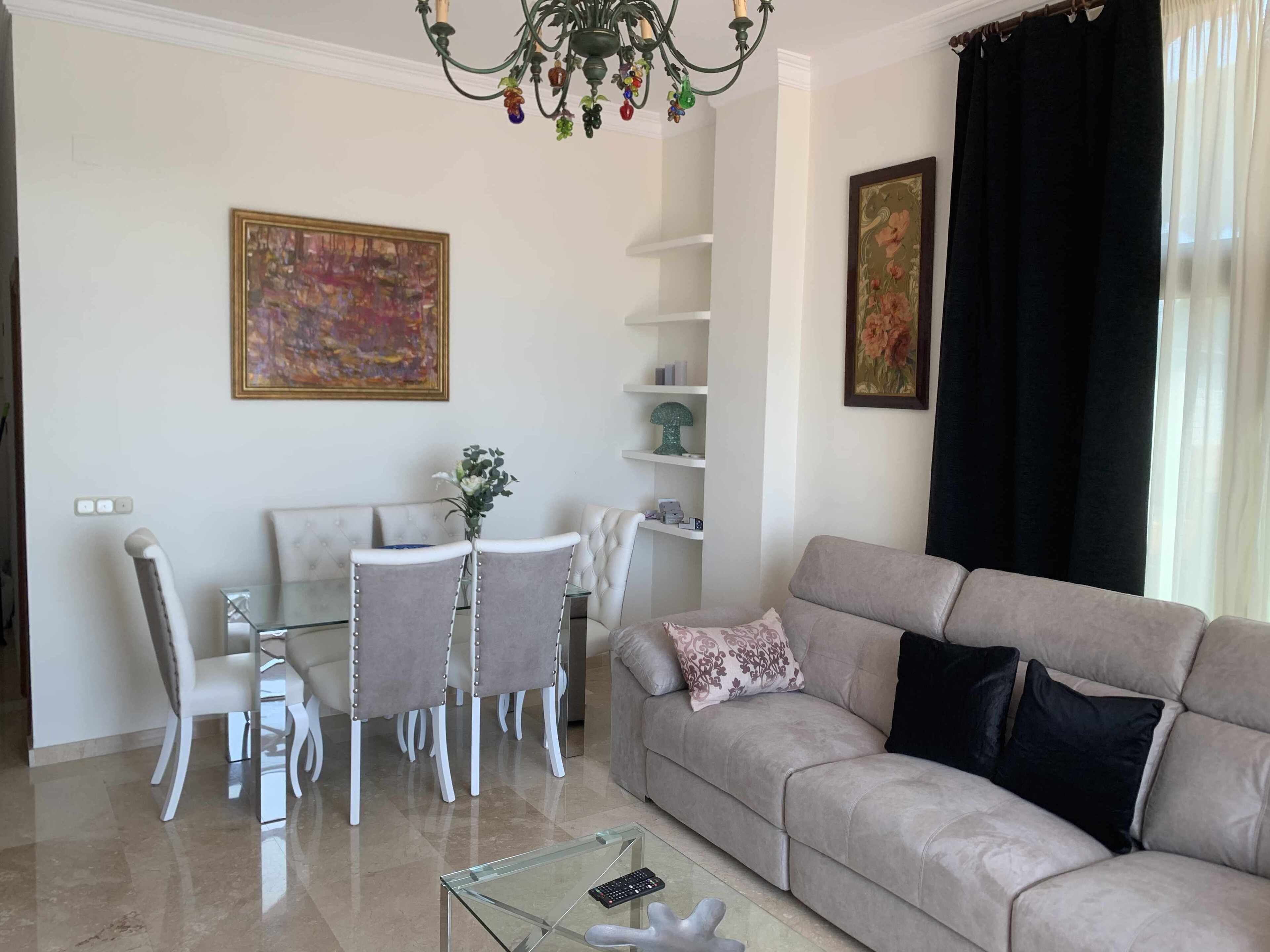 A living room with a beige sofa, a glass coffee table, a dining table with white chairs, and a chandelier.