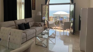 Un salon comprenant un canapé, des fauteuils, une table basse en verre et une vue sur la mer depuis un balcon ouvert.