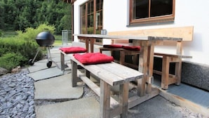 Een terras met houten banken en rode kussens, een barbecue en een wit gebouw met grote ramen.