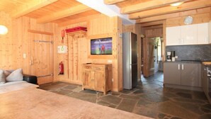 Een houten interieur met een stenen vloer, een aan de muur bevestigde televisie en een keuken met kasten en apparatuur.