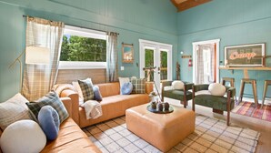 Un salon confortable avec un canapé, un pouf et un fauteuil, un coin repas avec une table et des chaises, et une pancarte indiquant « Camp ».