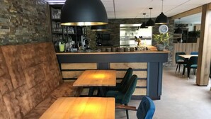 Ein gemütliches Café mit Holztischen, gepolsterten Sitzgelegenheiten, einer Bar und einer sichtbaren Küche im Hintergrund.