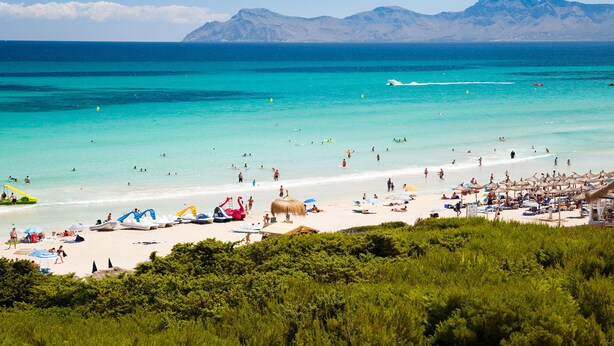 A beach with clear turquoise waters, a sandy shore, and mountains in the background.