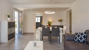 A modern living room with a sofa, a dining table with chairs, a kitchen in the background, and a large window.
