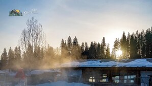 Ein Thermalbecken mit aufsteigendem Dampf, umgeben von schneebedeckten Bäumen und einem Holzhaus mit rotem Dach.